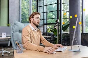 Young serious thinking businessman at work inside office with computer, blond man in casual clothes at desk thinking about tasks and ideas. photo