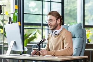 Young handsome male blogger sitting at the desk in the office at the computer in front of the microphone. Records a podcast, talks on a call, communicates in an online chat. photo