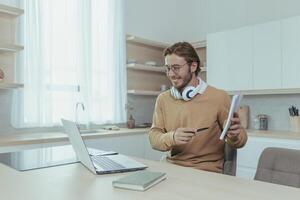 Young teacher teaches remotely from home, man works in kitchen at home, uses laptop for online call with communicates with students smiling explains assignment shows notebook. photo