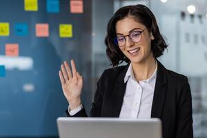 Businesswoman waving at the camera during a call in a modern office, representing remote work and virtual communication. photo