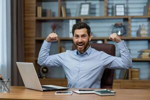 Ecstatic bearded male in blue shirt raising hands with clenched fists in winner pose while working by computer. Joyful entrepreneur feeling rejoiced while screaming and celebrating successful day. photo