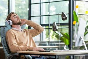 Young businessman man resting from work, overtime, break. Sitting in the office at a table with a computer, leaning back in a chair, listening to music in headphones, closed his eyes, relaxed.. photo