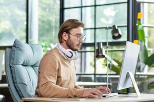 Young serious male student in headphones, beige sweater and glasses. He sits at the table, concentrates on studying at the computer. Writes on the keyboard, talks on a call, listens to lectures. photo