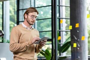 Young successful thinking programmer inside loft style office uses tablet computer, young blond man in casual clothes reads news online, serious businessman browses internet pages. photo