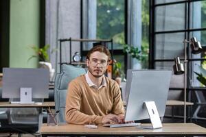 Portrait of thinking serious man with computer inside modern green loft office, blond man looking thoughtfully at camera businessman in casual clothes working with computer. photo