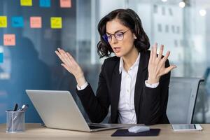 Angry woman in business attire expressing frustration while working on laptop in modern office setting. Post-it notes visible on the glass wall in background. photo