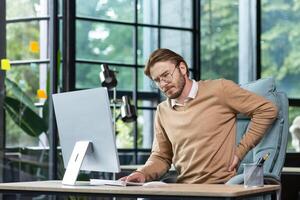 Tired businessman at workplace has severe back and lower back pain, young man working sitting at table with laptop and computer in middle of office. photo