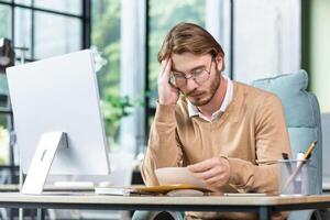 Man upset in office reading message on paper, businessman received mail envelope with notification and bad news, sitting at workplace with computer. photo