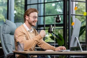 Young successful entrepreneur working inside office with computer, man in casual clothes smiling and typing on keyboard, businessman happy with achievement results and income. photo