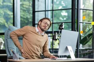 Tired businessman at workplace has severe back and lower back pain, young man working sitting at table with laptop and computer in middle of office. photo