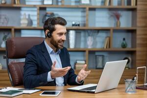 Positive man in wireless headset with micro hosting conference while sitting by desktop with computer. Millennial businessperson using modern technology for online communication with partners. photo