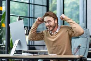 Young man in office celebrating victory and successful achievement results, businessman holding hands in triumph gesture and reading good news from computer , blond programmer in casual clothes. photo