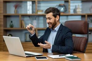 Online operation of buying and selling real estate. Young businessman realtor sits in the office and talks on a call with clients, shows the keys to the camera and offers product options. photo