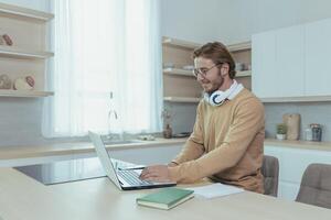 Young successful and happy businessman working from home, man using laptop for remote work, freelancer smiling in casual clothes sitting in kitchen at table. photo