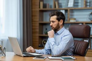 Focused bearded man looking attentively at portable computer screen and holding hand under chin in workspace. Small business owner checking monthly statements online and accounting bills. photo