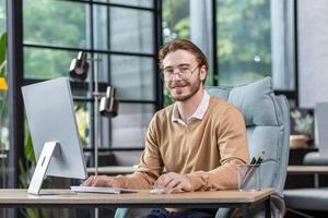 Portrait of successful smiling programmer inside modern green loft office, blond man smiling and looking at camera, businessman in sweater and casual shirt close up working with computer. photo