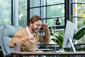 Young man in office celebrating victory and successful achievement results, businessman holding hands in triumph gesture and reading good news from computer , blond programmer in casual clothes. photo