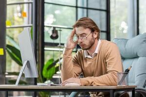 Problems at work. A young man, a specialist. office desk, an accountant sits in the office at a table with a computer. Holds his head, thinks, worries. photo