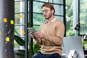 Young successful thinking programmer inside loft style office uses tablet computer, young blond man in casual clothes reads news online, serious businessman browses internet pages. photo