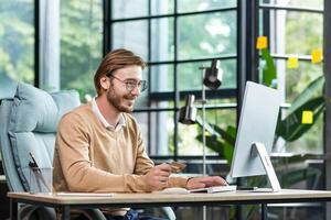 Online transactions with a credit card. A smiling young man sits in the office at the table, looks at the computer, holds a credit card in his hands. Successful transfer, purchase, order. photo