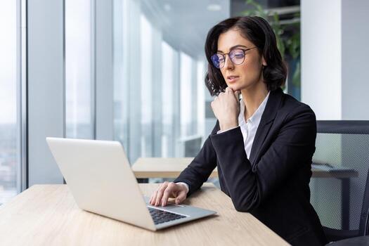 grave mujer de negocios jefe pensando trabajando con ordenador portátil dentro oficina cerca ventana, hembra oficina trabajador en negocio traje mecanografía ideas en teclado, mirando a pantalla leyendo y resolviendo Actual Tareas foto