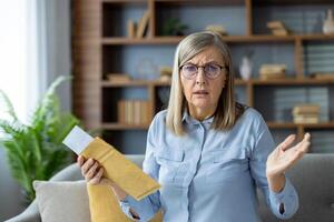 Confused senior woman holding an envelope with a questioning expression while sitting in a living room setting. photo