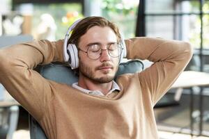 Portrait of a tired office worker in headphones resting at the workplace in a chair, arms crossed on the title with closed eyes, napping during a break between work, listening to music. photo