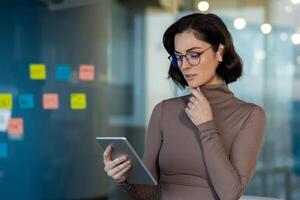 Young thinking woman with tablet computer in hands at workplace inside office, Businesswoman standing near window using app, deciding and brainstorming startup idea. photo