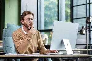 Portrait of a young designer, architect, engineer sitting in a modern office, working at a computer. He holds his chin with his hand, looks intently and thoughtfully at the monitor. photo