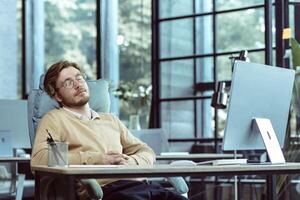 Tired IT specialist engineer programmer sleeps in the office at the table during the day, man in glasses and casual clothes works at the computer, businessman rests photo