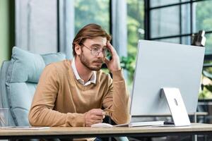 Credit card transaction failed. An upset young man sits in the office at the table, looks at the computer, holds a credit card in his hand. Disappointed, he leaned on his arm. photo