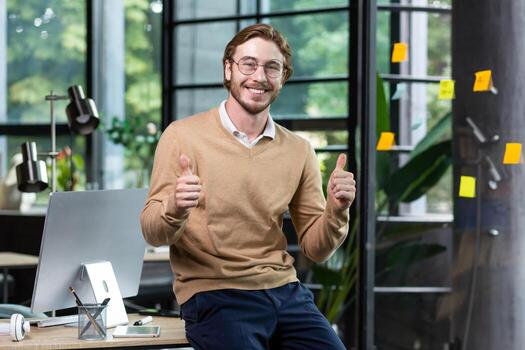 Portrait of happy blond man inside office, man in casual clothes smiling and looking at camera, young successful businessman showing thumbs up at camera, programmer at work with computer. photo