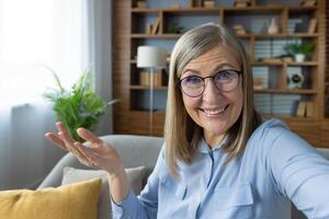 Smiling woman with glasses chatting on a call while waving. Home office in the background. Joyful and friendly communication. photo