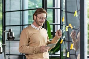 Smiling young man businessman, programmer, freelancer standing in the office, looking at a tablet, drawing a progress graph on a transparent board. Glue colored papers, notes, stickers, hints. photo