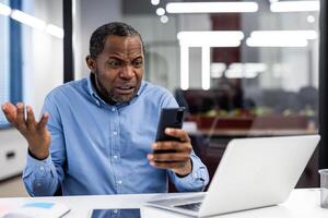 Confused office worker holding phone with puzzled expression at desk with laptop and tablet. Image depicts frustration, confusion, and technology issues in modern business environment. photo
