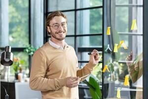 A young designer, programmer, freelancer man is standing in the office, holding a tablet, drawing a progress graph on a transparent board. Glue colored, stickers, hints. He looks at the camera, smiles. photo