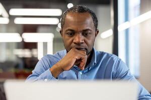 Professional businessman concentrating on tasks using laptop in modern office environment. Serious expression reflects deep thought. Ideal for themes emphasizing business, technology, productivity photo