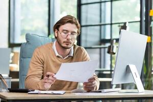 A serious young male programmer sits in the office at a table, works at a computer and documents. He holds a project, a plan, bills in his hands. photo