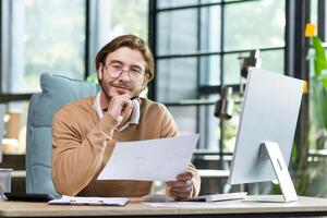 Portrait of a young designer, architect, engineer sitting in an office, working at a computer. He holds chin with his hand, holds documents, a plan, a project in his hands. He smiles at the camera. photo