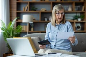 Senior woman looking worried while managing finances at home, using a laptop and holding documents. Concept of financial stress, budgeting, and retirement planning. photo