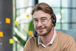 Portrait of young man with headset for call inside office, close-up tech support worker in glasses smiling and looking at camera, businessman blond customer service online help. photo