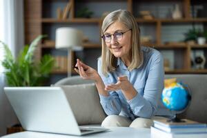 Senior woman teacher conducting an online class from her home, explaining with gestures while looking at a laptop. The setup includes educational materials indicating a virtual teaching environment. photo