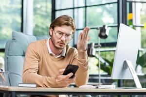 Upset young male programmer, freelancer sitting in the office at the table, holding the phone in his hands, looking worriedly. There was a problem, bad news, messages, checking the account. photo
