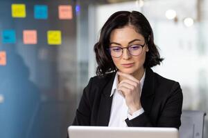 Thoughtful entrepreneur in formalwear resting head on hand while looking attentively at portable computer screen. Concentrated female checking accounting reports with bills with basic expenses. photo
