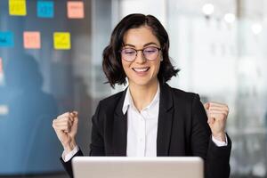 Successful female in formal suit doing yes gesture with clenched fist while looking at laptop screen on blurred background. Smiley accountant getting positive email from head of department in office. photo