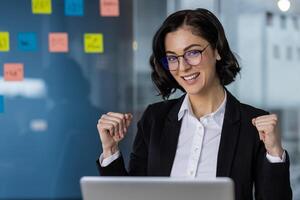 Happy businesswoman in formal attire celebrating a success with fists raised, working on laptop in modern office. photo
