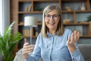 Senior woman with glasses engaging in a call while looking and talking to the camera. She seated in a cozy living room with shelves and plants in the background, smiling warmly and gesturing. photo