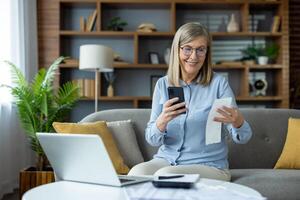 Senior woman sitting on couch using smartphone and computer while checking receipts. Concept of remote work, home finance management, and online accounting. Modern cozy home interior. photo
