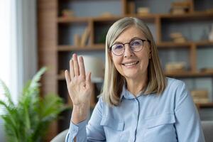 Senior woman with glasses smiling and waving while engaging in a call from home, representing communication and connection. photo