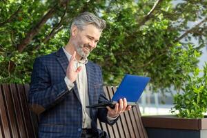 Affable senior businessman with a tablet engaging in a call waves hello from a bench outside an office building. photo
